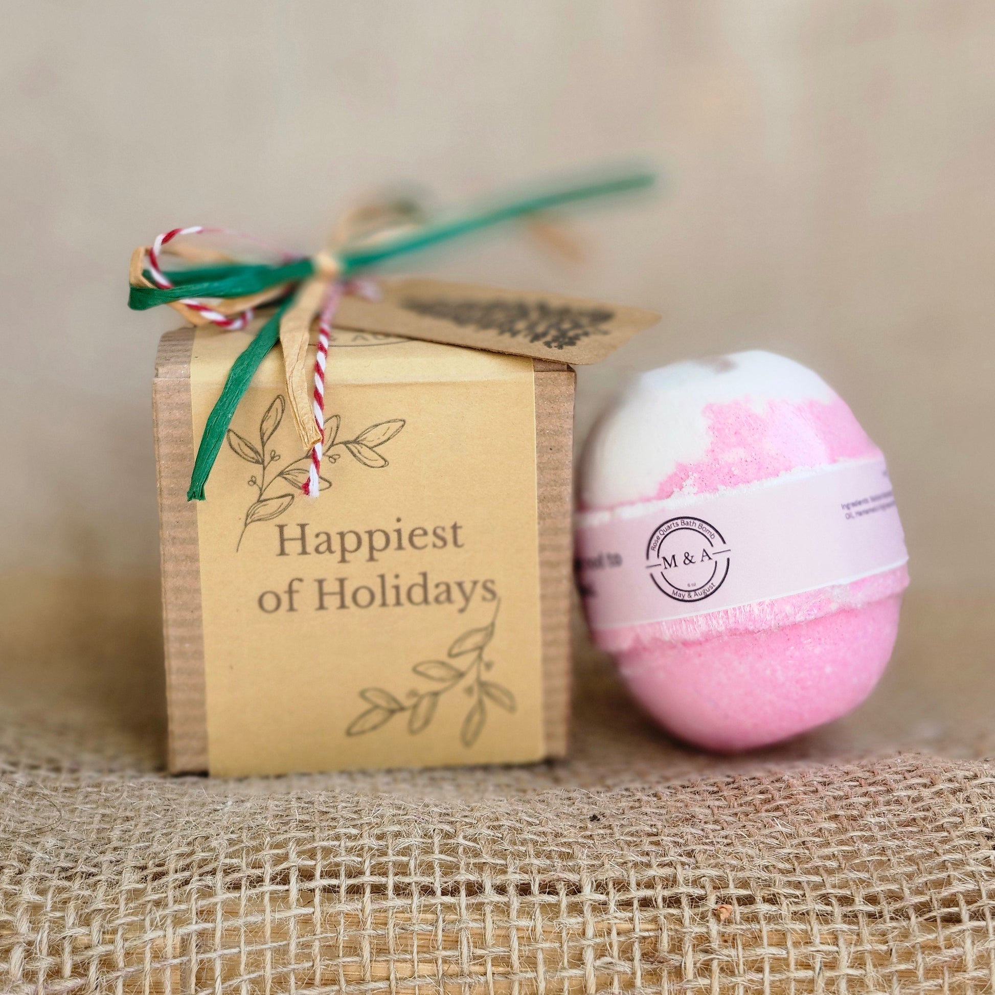 Pink bath bomb with a label on top of a wooden box labeled 'Happiest Holidays' against a textured beige background.