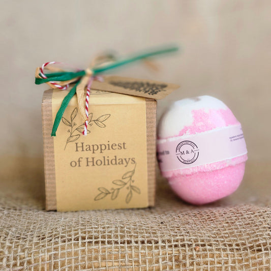 Pink bath bomb with a label on top of a wooden box labeled 'Happiest Holidays' against a textured beige background.